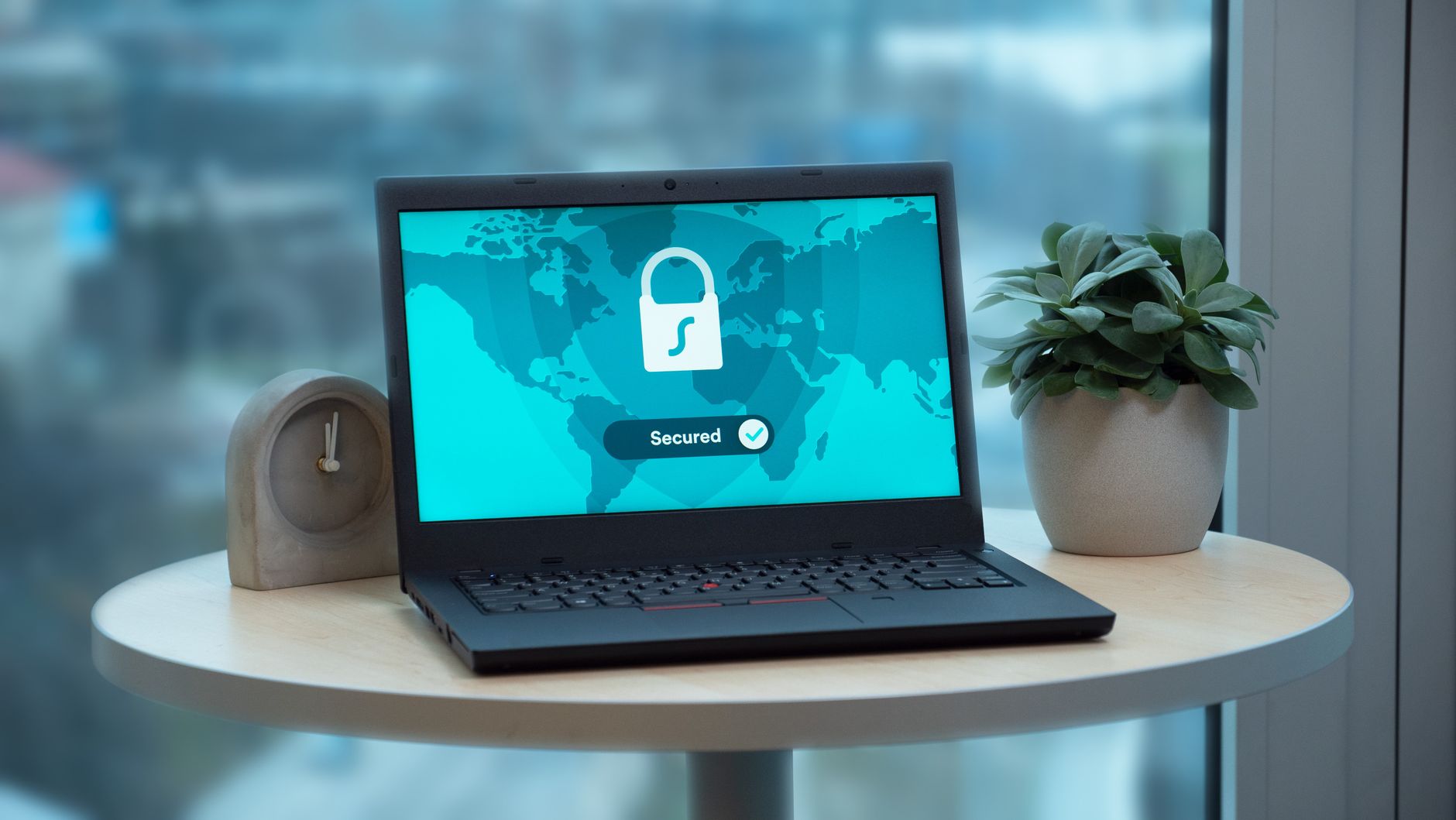 Laptop displaying a secured lock icon with global map and checkmark on screen, accompanied by a desktop clock and potted plant on a round table