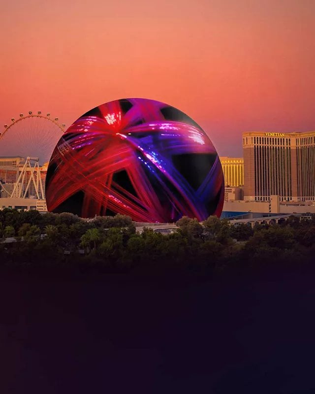 Large illuminated red and purple spherical structure at dusk with cityscape and observation wheel in background