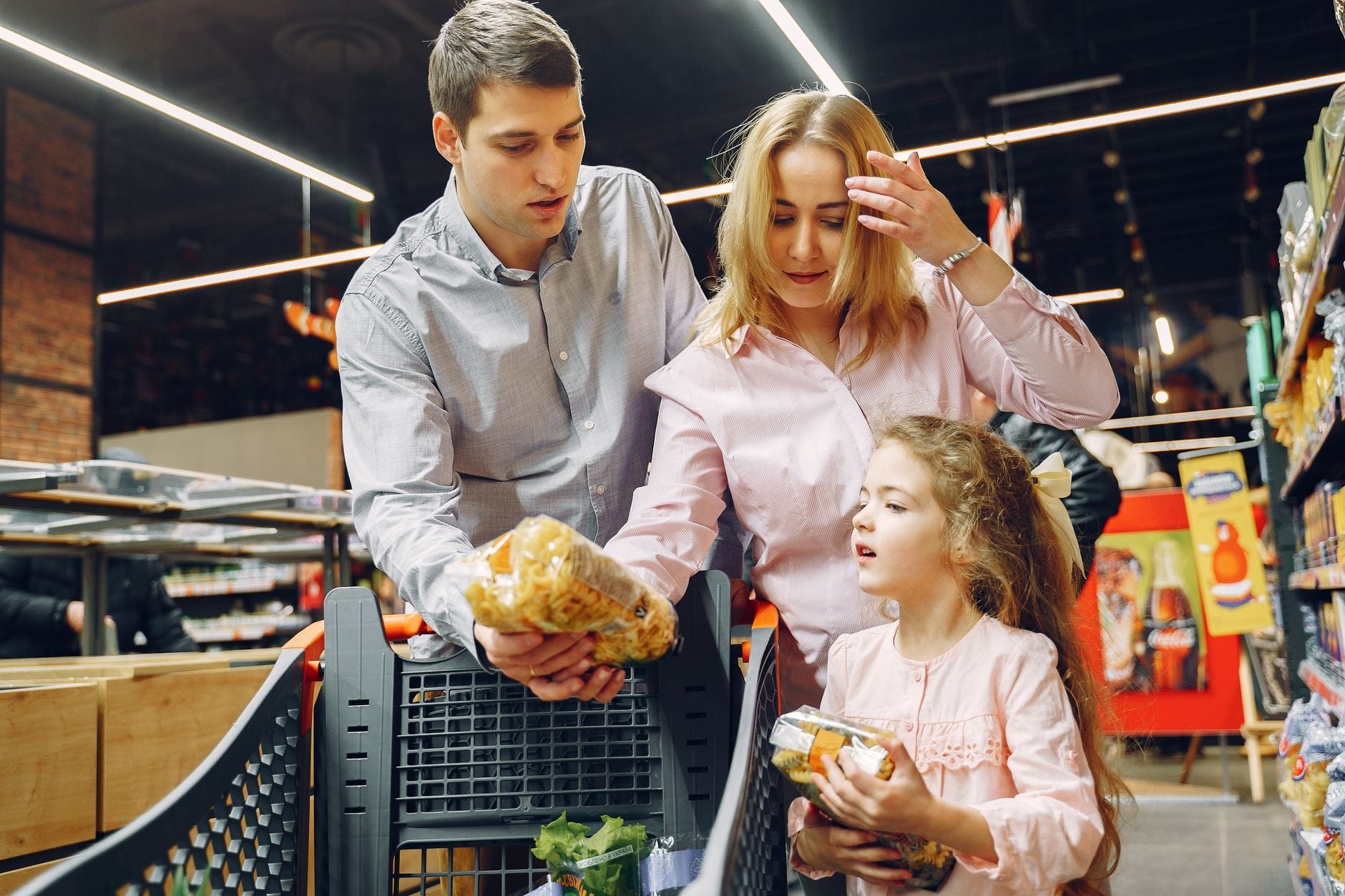 Family of three shopping in grocery store aisle, examining packaged food items in cart