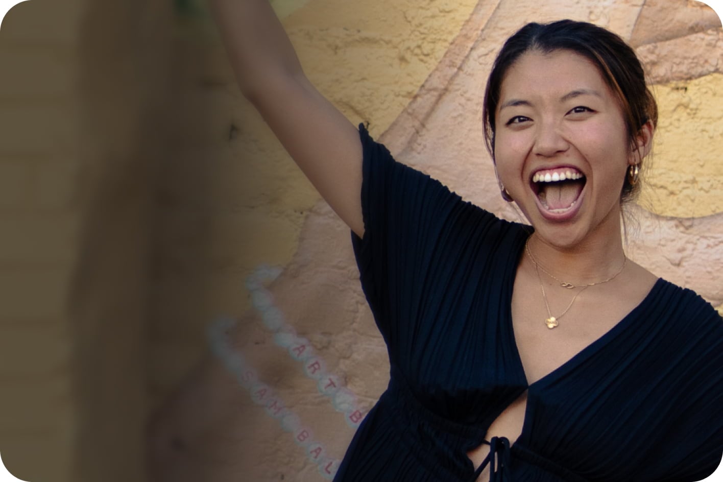 Woman smiling with arm raised against yellow brick wall wearing black ribbed top