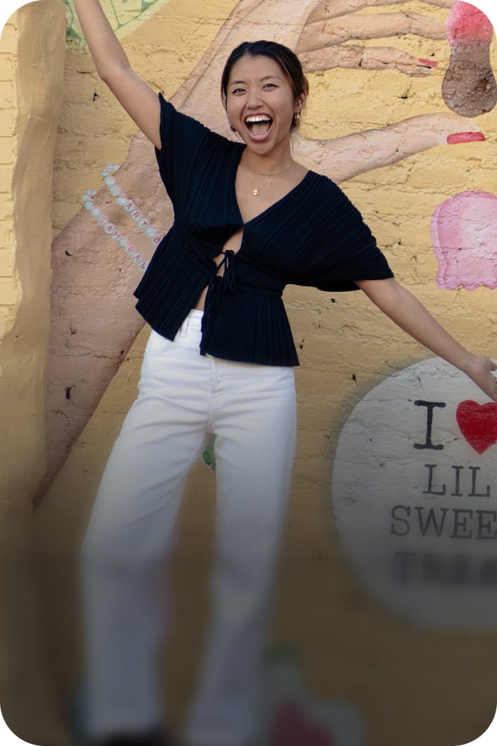 Woman laughing with arm raised against yellow brick wall wearing black top and white pants